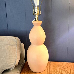 May include: A peach-colored gourd-shaped table lamp with a brass neck and a white lightbulb. The lamp sits on a wooden table next to a gray couch. The background is a dark blue wall.