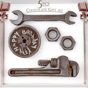 May include: A chocolate gift set featuring tools like a wrench, gear, and nuts. The gear has "HAPPY BIRTHDAY ALEX" written on it. The set includes chocolate bars and is presented in a white box.