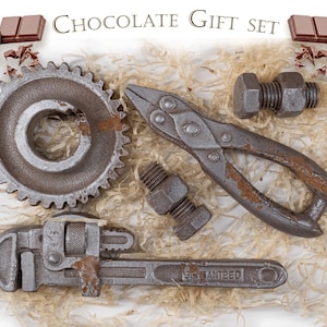 Pode incluir: Um conjunto de presente de chocolate com ferramentas, incluindo uma engrenagem, alicate, chave inglesa e parafusos. O conjunto inclui barras de chocolate e é apresentado em palha. O texto "Chocolate Gift Set" está no topo.