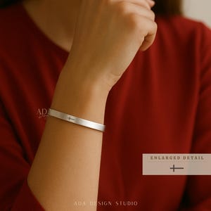 May include: A silver-colored cuff bracelet with a cross symbol engraved on the front. The bracelet is shown on a person's wrist, with the text "PERSONALIZED BRACELET" at the top and "ADA DESIGN STUDIO" at the bottom. The enlarged detail of the cross is shown on the right.