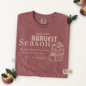 May include: A brick-colored t-shirt with the words "apple pickin' HARVEST Season" printed in a light beige color. The shirt also features a graphic of a basket of apples and the text "The land yields its harvest: God, our God, blesses us!"