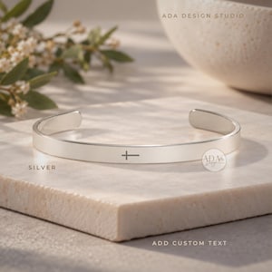 May include: A silver cuff bracelet with a smooth, polished finish and a small cross symbol engraved on the front. The text ADA DESIGN STUDIO is visible in the upper right, and the word SILVER is printed on the left with ADD CUSTOM TEXT below.