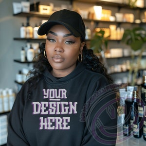 Puede incluir: Una persona lleva una sudadera con capucha negra con el texto "YOUR DESIGN HERE" en morado. Lleva una gorra de béisbol negra y tiene el pelo oscuro.