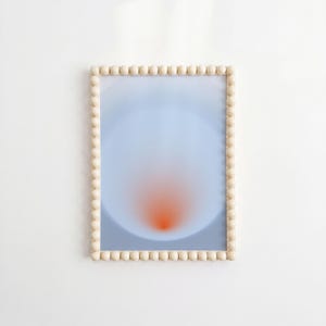 Può includere: Una stampa artistica rettangolare da parete caratterizzata da una sfumatura tenue di azzurro che transita verso un centro arancione vibrante, racchiusa in una cornice in legno chiaro decorata con un bordo continuo di piccole sfere di legno.
