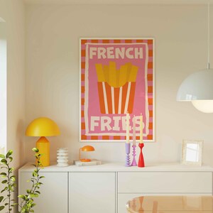 May include: A framed poster with the words "FRENCH FRIES" in pink and white, featuring a graphic of fries in a red container. A yellow lamp, candles, and other decor sit on a white cabinet.