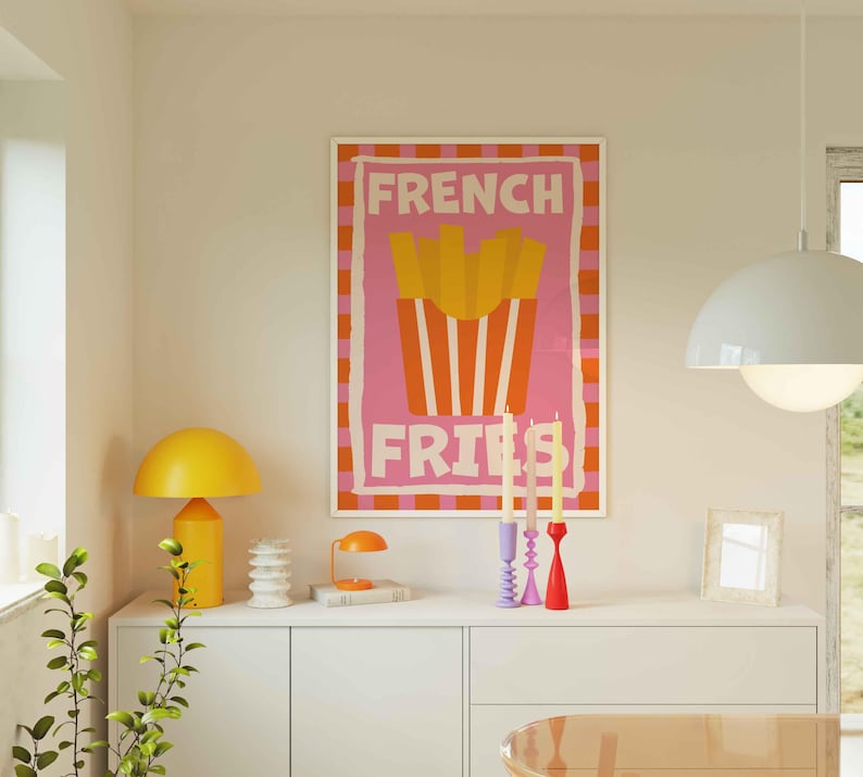 May include: A framed poster with the words "FRENCH FRIES" in pink and white, featuring a graphic of fries in a red container. A yellow lamp, candles, and other decor sit on a white cabinet.