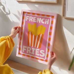 May include: Pink framed art print with the words "FRENCH FRIES" in white. The artwork features a pink background with a graphic of french fries in a red and white striped container. The frame is pink.