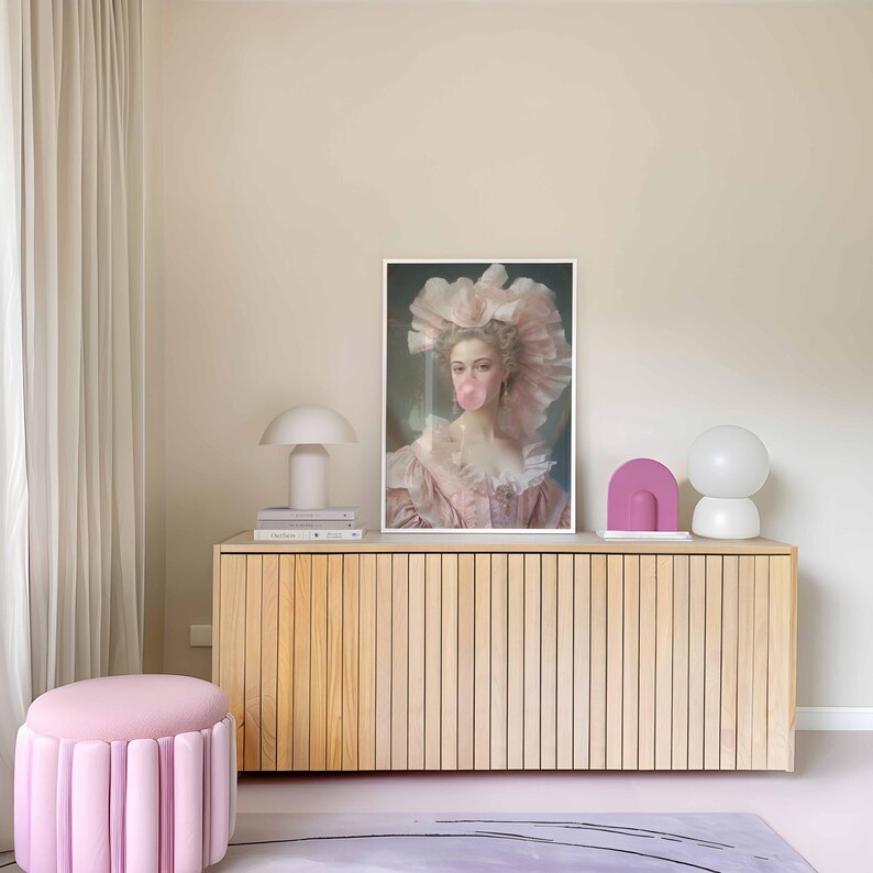 May include: A framed print of a woman in a pink dress blowing a bubblegum bubble, displayed on a light wood cabinet. The room features a pink ottoman, a white mushroom lamp, and decorative objects, creating a soft, pastel aesthetic.