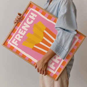 May include: A framed art print featuring a pink background with the words "FRENCH FRY" in white. The artwork depicts a stylized image of golden french fries. The frame is a light wood color with orange and white stripes.