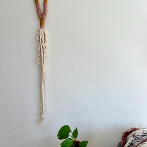 以下が含まれることがあります： マスタードイエロー、ピンク、クリーム色の手作りのマクラメ壁掛け。 上部のループは糸で包まれ、結び目のフリンジに変わります。 緑の葉を持つ鉢植えの植物が下にあります。 約100cmの長さです。