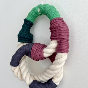 Może przedstawiać: Ręcznie wykonana, abstrakcyjna dekoracja ścienna. Element wykonany jest z grubej, skręconej liny w kształcie ósemki. Sekcje owinięte są zieloną, ciemnozieloną, bordową, granatową i kremową przędzą. Mały metalowy pierścień u góry do zawieszenia.