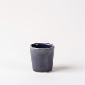 May include: A small, dark blue ceramic cup with a slightly tapered shape. The interior of the cup is a deeper shade of blue. The cup is set against a plain white background.