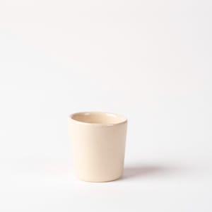 May include: A small, cream-coloured ceramic cup with a cylindrical shape. The cup has a smooth, matte finish and a slightly rounded rim. It is empty and sits on a white background. The cup is likely used for drinking beverages.