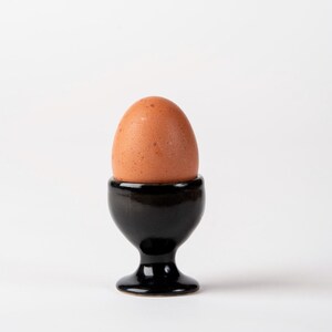 Puede incluir: Un huevo marrón en un huevera de cerámica negra. La huevera tiene una base de pedestal y una parte superior redondeada. El fondo es blanco.
