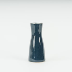 May include: A small, dark teal ceramic vase with a glossy finish. The vase has a narrow neck and a wider base, with a slightly flared opening. The vase is approximately 15 cm tall.