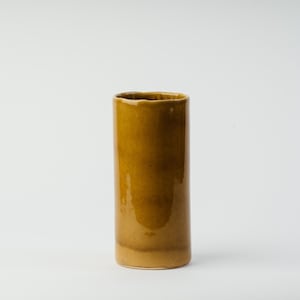 May include: A tall, cylindrical ceramic vase in a warm, mustard yellow hue. The vase has a slightly textured surface and a gently uneven rim. The design is simple and modern, suitable for displaying flowers or as a decorative accent.