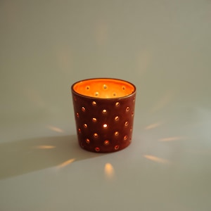 Puede incluir: Un portavelas de terracota con un patrón de puntos. El portavelas está encendido y la luz brilla a través de los agujeros.