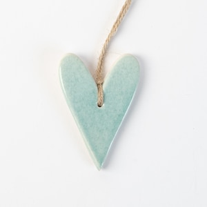 May include: A pale blue ceramic heart-shaped decoration with a glossy finish. It is suspended by a natural twine string, ideal for hanging. The ornament is set against a clean white background.