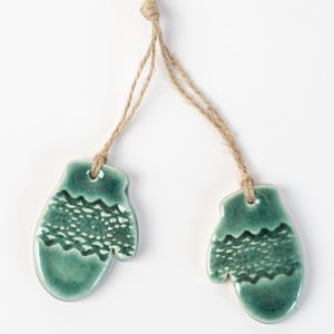 Puede incluir: Dos adornos de cerámica con forma de mitones en color verde turquesa, cada uno con un diseño texturizado y una cuerda de yute para colgar. Los mitones son una decoración con temática invernal, perfecta para un árbol de Navidad.