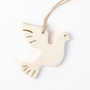 May include: A cream ceramic dove ornament with brown detailing on the wings and tail. It has a natural twine loop for hanging. The dove is a symbol of peace, suitable for home decoration.