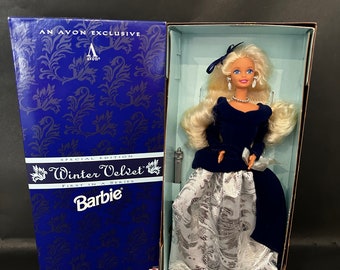Può includere: Una bambola Barbie blu e argento con un abito di velluto con un bordo di pizzo bianco. La bambola è in una scatola con le parole "Winter Velvet" e "First in a Series" su di essa. La scatola è blu con un motivo argentato.