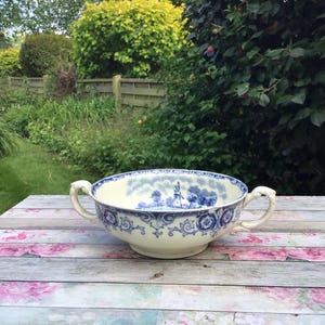 May include: A cream-coloured ceramic bowl with blue floral and landscape designs. The bowl has two ornate handles and a wide rim. The interior features a blue landscape scene with a windmill. It sits on a floral patterned surface.