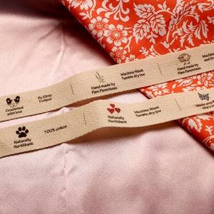 May include: Close-up of a beige cotton fabric label with brown text and icons. The label reads "Crocheted with love", "By Elroy Trollpon", "Hand made by Pam Parenteau", "Machine Wash Tumble dry low", "Naturally Northbank", "100% cotton", and "Naturally Northbank".