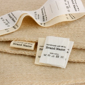 K&ouml;nnte beinhalten: Wei&szlig;e Baumwollstoff-Etiketten mit schwarzem Text, der "Handmade with &hearts; by Brand Name" lautet und Pflegehinweise enth&auml;lt. Die Etiketten sind gefaltet und auf einer beigefarbenen Stoffoberfl&auml;che gestapelt.