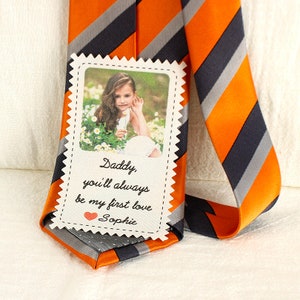 May include: A white card with a photo of a young girl and the text "Daddy, you'll always be my first love  ❤️ Sophie" attached to a striped orange and black tie.