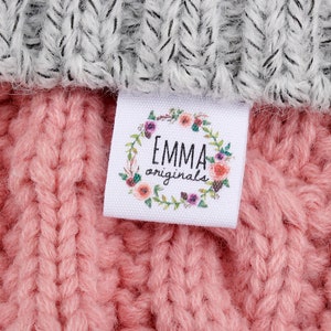 May include: A white label with a floral wreath and the text "EMMA originals" in black lettering. The label is attached to a pink knitted fabric.