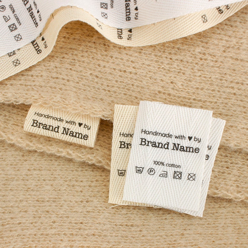 K&ouml;nnte beinhalten: Zwei wei&szlig;e Stoff-Etiketten mit schwarzem Text. Das obere Etikett lautet "Handmade with &hearts; by Brand Name 100% cotton". Das untere Etikett lautet "Handmade with &hearts; by Brand Name".