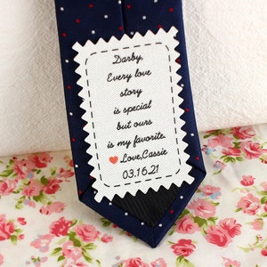 May include: A white tag with a scalloped edge and black stitching, attached to a blue tie with white and red polka dots. The tag has a handwritten message that reads "Darby, Every love story is special but ours is my favourite. Love, Cassie 03.16.21".