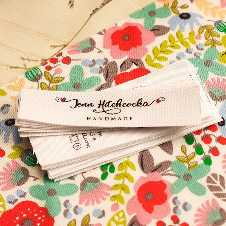 Puede incluir: Una pila de etiquetas de tela blanca con el texto "Jenn Hitchcocka HANDMADE" en escritura cursiva negra. Las etiquetas est&aacute;n sobre un fondo blanco con un patr&oacute;n floral en rojo, rosa, verde y azul.