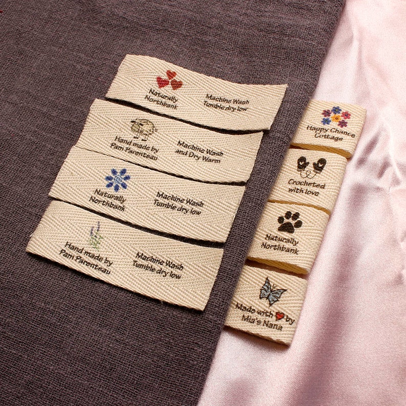 May include: A collection of eight beige fabric labels with various designs and text. The labels include a heart, a sheep, a flower, a lavender sprig, a paw print, a butterfly, a pair of mittens, and a floral design. The text on the labels includes "Naturally Northbank", "Hand made by Pam Parenteau", "Machine Wash Tumble dry low", "Machine Wash and Dry Warm", "Crocheted with love", "Happy Chance Cottage", and "Made with by Mia's Nana".