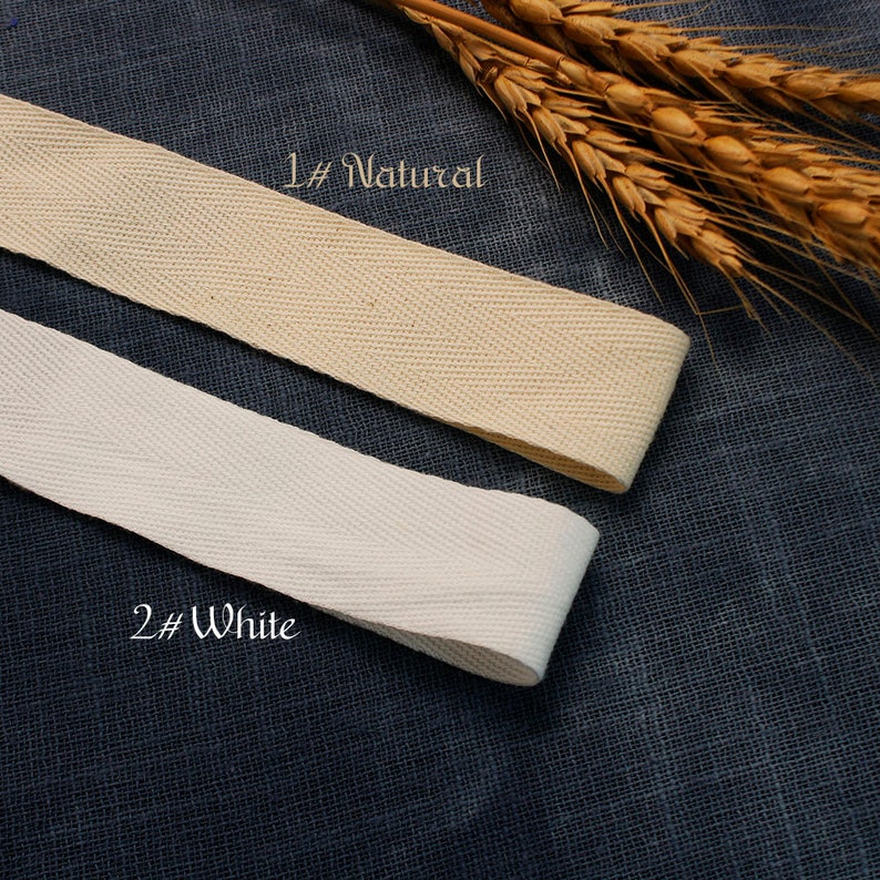 May include: Two samples of cotton twill tape, one natural and one white. The tape is woven with a herringbone pattern. The tape is displayed on a textured fabric background.