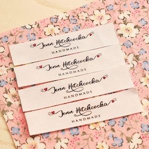 Puede incluir: Cuatro etiquetas de tela blanca con el texto "Jenn Hitchcocka Handmade" en letras cursivas negras con un s&iacute;mbolo de coraz&oacute;n rojo antes del nombre. Las etiquetas est&aacute;n apiladas una encima de la otra sobre un fondo de tela floral rosa.