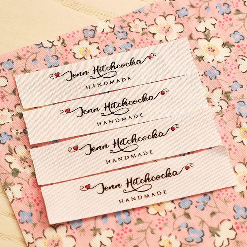 Puede incluir: Cuatro etiquetas de tela blanca con el texto "Jenn Hitchcocka Handmade" en letras cursivas negras con un s&iacute;mbolo de coraz&oacute;n rojo antes del nombre. Las etiquetas est&aacute;n apiladas una encima de la otra sobre un fondo de tela floral rosa.