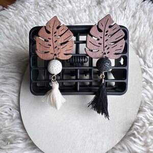 May include: Two car air vent diffusers. Each diffuser features a wooden leaf design, a decorative bead, and a tassel. One has a white bead and tassel, the other has a black bead and tassel. The diffusers are attached to a black vent clip.
