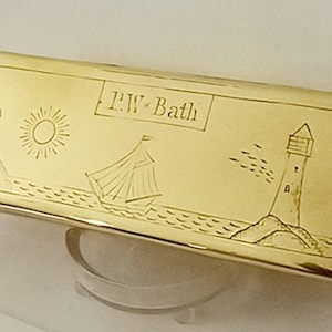 May include: A gold rectangular box with an engraved design of a sailboat, lighthouse, sun, and trees. The box is inscribed with the text "P.W. Bath".