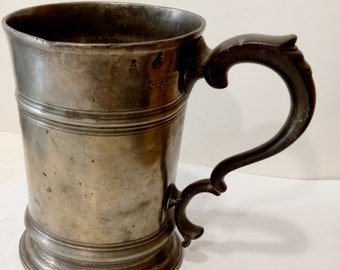 May include: A pewter tankard with a dark brown handle. The tankard has a cylindrical body with a flared base and a lip. The handle is ornate and has a scroll design. The tankard is marked with a maker's mark.