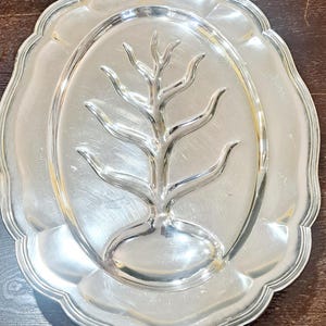 May include: A silver serving platter with a tree design in the center. The platter has a scalloped edge and is oval shaped.