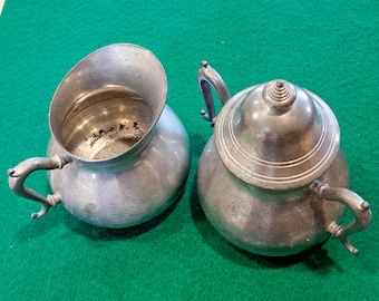 May include: A vintage pewter creamer and sugar set with ornate handles. The creamer has a wide, rounded body and a flared lip. The sugar bowl has a domed lid with a finial.