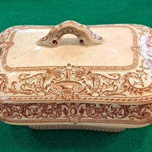 May include: A white ceramic serving dish with a brown floral design. The dish has a lid with a handle and two side handles.