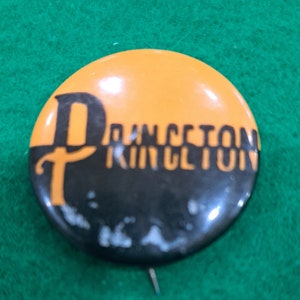 May include: A black and orange button with the word "Princeton" written in black letters.