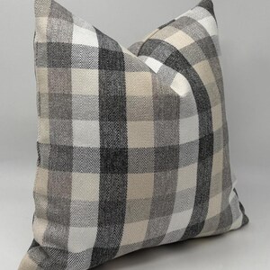 May include: Square decorative pillow with a plaid pattern in shades of gray, black, and cream. The pillow has a soft, textured appearance and a classic, neutral design. The pillow is a square shape.