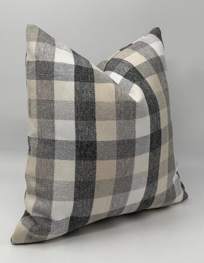 May include: Square decorative pillow with a plaid pattern in shades of gray, black, and cream. The pillow has a soft, textured appearance and a classic, neutral design. The pillow is a square shape.