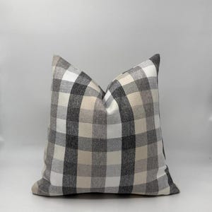 May include: A square decorative pillow with a classic buffalo check pattern. The pillow features a repeating grid of black, white, and beige squares. The pillow is a neutral color and would complement a variety of home decor styles.