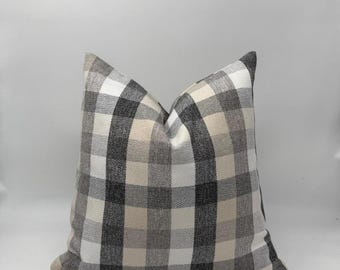 May include: A square decorative pillow with a classic buffalo check pattern. The pillow features a repeating grid of black, white, and beige squares. The pillow is a neutral color and would complement a variety of home decor styles.