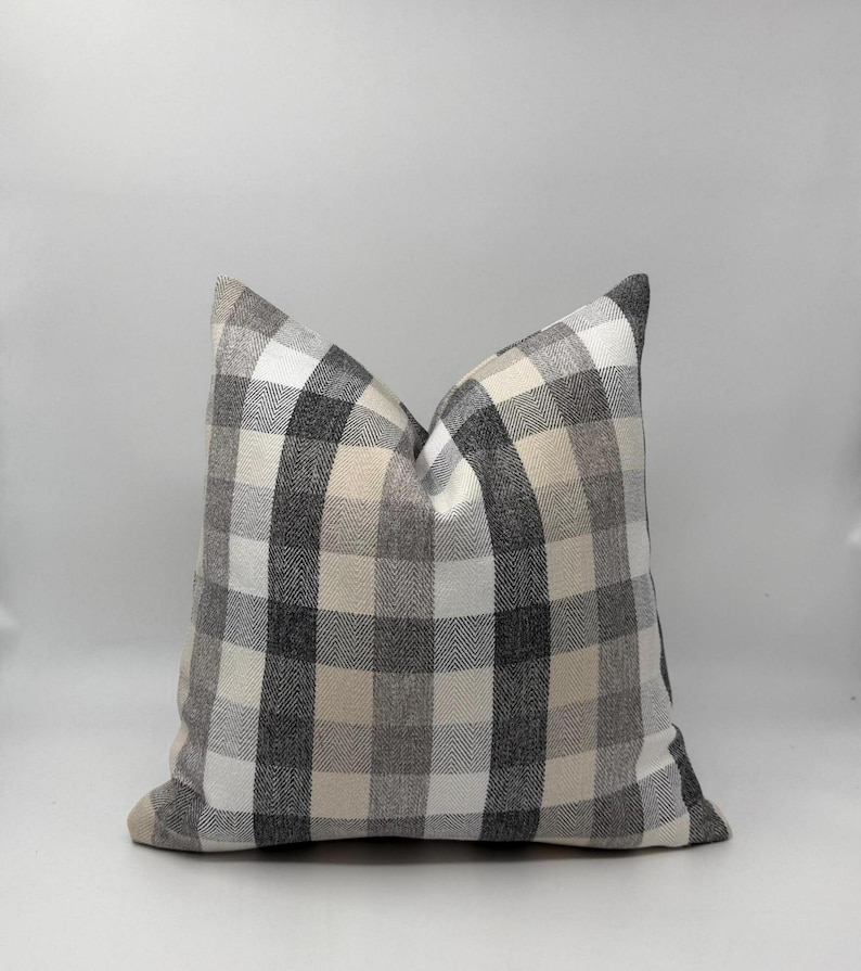 May include: A square decorative pillow with a classic buffalo check pattern. The pillow features a repeating grid of black, white, and beige squares. The pillow is a neutral color and would complement a variety of home decor styles.