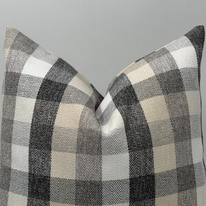May include: A decorative pillow with a large, checkered pattern in shades of gray, white, and beige. The pillow has a soft, textured appearance, with a classic, neutral design. The pillow is a square shape.